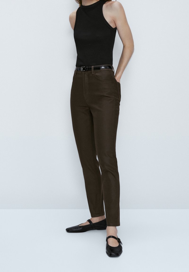 Zwarte mouwloze top, bruine slim-fit broek en zwarte flats. De broek heeft zijzakken en een gladde textuur met een riem bij de taille.