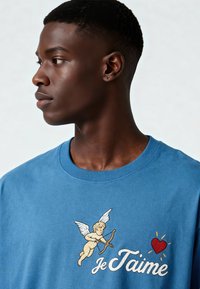 T-shirt en coton bleu avec un chérubin brodé aux ailes déployées, tenant un arc, accompagné du texte « Je T'aime » et d'un dessin de cœur rouge.