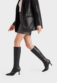 Stivali neri al ginocchio con punta affusolata e design elegante. Realizzati in pelle liscia e dotati di una zip laterale per una facile vestibilità.