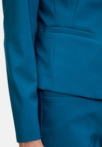 Zöldeskék szabott öltöny, strukturált blézerrel és testhezálló nadrággal. A blézer rejtett zsebbel és sima textúrával rendelkezik.