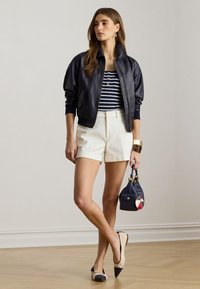 Jachetă bomber din piele neagră peste un top cu dungi albastre și albe, asortată cu șorturi crem și balerini bicolori, ținând o geantă de mână bleumarin.