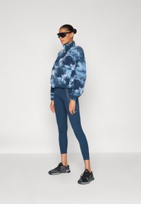Blue tie-dye fleece pullover with high collar and front pocket, paired with solid dark blue leggings. Black athletic shoes complete the outfit.