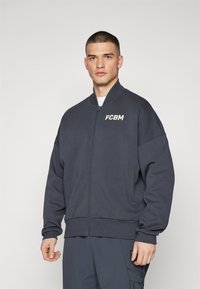 Dunkelgraue, mit Reißverschluss versehene Bomberjacke mit gerippten Bündchen und Saum. Verfügt über das „FCBM“-Logo auf der linken Brust. Der Stoff ist weich und hat eine lockere Passform.