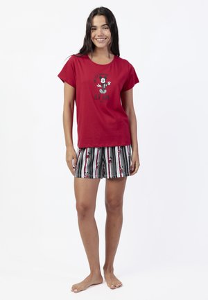 Joven sonriendo, vistiendo una camiseta roja de Mickey Mouse y pantalones cortos a rayas en blanco y negro con estampado de Mickey Mouse, de pie y descalza.