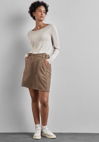 Maglia a maniche lunghe in maglia beige chiaro abbinata a una mini gonna brown con cintura e grandi tasche frontali. Il modello indossa sneakers bianche.