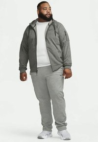 Grå Nike hettejakke med glidelås, med en hvit skjorte under og grå joggebukser. Har en stofftekstur med lommer og en avslappet passform.