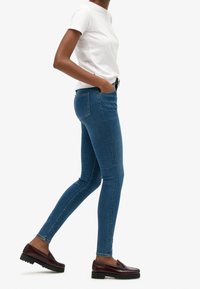 Vaqueros ajustados de color azul con una textura suave y diseño de cinco bolsillos, combinados con una camiseta blanca sencilla y mocasines oscuros.