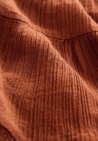 Rudos spalvos audinys su tekstūruotomis vertikaliomis juostomis, minkšta megzta medžiaga ir subtili nelygumų pynė, suteikianti gilumo.