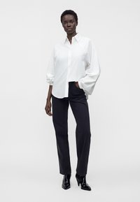 Femme portant une chemise blanche ample boutonnée avec des manches ballon, un pantalon noir droit et des bottines noires pointues sur fond uni.