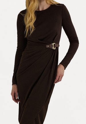 Vrouw draagt een donkerbruine jurk met lange mouwen, met een zijgesp detail en geplooide stof ter hoogte van de taille.