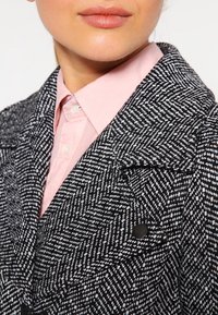 Abrigo de pata de gallo en blanco y negro con cuello muesca, detalle de botones y tejido texturizado, llevado sobre una camisa de cuello rosa claro.