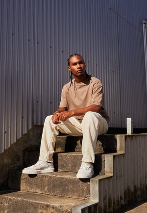 Beige t-shirt, avslappnade krämfärgade byxor och vita sneakers. Sitter på betongtrappor, mot en texturerad metallbakgrund.