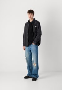 Black denim shirt over a black knit top, paired with light blue distressed wide-leg jeans and black shoes. Model stands against a plain background.