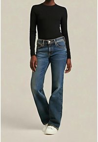 Personne portant un jean évasé bleu marine, un haut à manches longues noir et des baskets blanches, debout devant un fond uni.