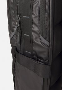 Gros plan sur le côté d'un sac à dos noir montrant une fermeture éclair avec une languette de tirage et une large sangle en tissu avec des boucles cousues.