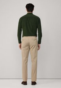 Ciemnozielona koszula z długim rękawem połączona z jasnobeżowymi spodniami, posiadającymi tylnie kieszenie i gładką teksturę. Buty są ciemnobrązowe.