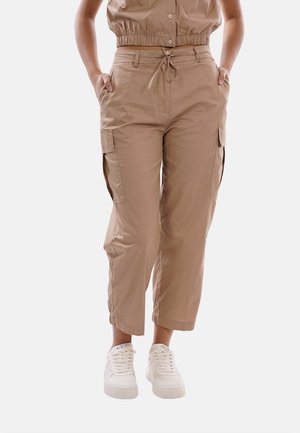 Persona che indossa pantaloni cargo beige corti con tasche laterali e sneakers bianche, in piedi con le mani nelle tasche.