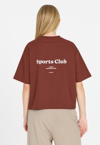 Kvinna med långt blont hår som bär en lös maroon T-shirt med texten "Sports Club Luxury Athleisure Wear LES ARCS" på ryggen.