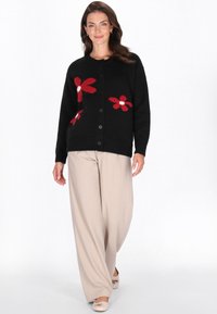 Cardigan nero con motivi floreali rossi, scollo rotondo e chiusura a bottoni. Abbinato a pantaloni beige a gamba larga e scarpe di colore chiaro.