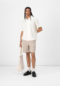 Hvid skjorte med korte ærmer og struktureret mønster, beige stribede shorts, sorte loafers og en lys tote bag. Enkle, neutrale farver.