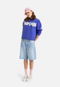 Sudadera azul con estampado floral blanco, combinada con shorts de mezclilla claros. Los accesorios incluyen una gorra rosa y zapatillas blancas con calcetines rosas.