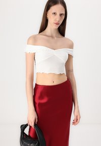 Biely crop top bez ramienok s volánmi, skombinovaný s červenou saténovou sukňou. Drží čiernu kabelku s oblým úchytom.