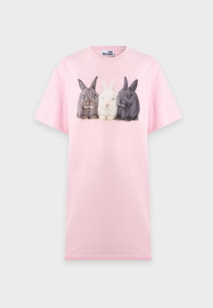 Femme portant une robe rose avec un imprimé de trois lapins sur le devant et des sandales noires à talons ouverts, se tenant devant un fond blanc uni.