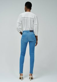 Blusa blanca ligera con detalle de encaje, combinada con jeans ajustados azules. La modelo lleva tacones beige, mostrando un look casual desde atrás.