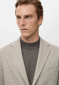 Hellgrauer Blazer mit strukturierter Oberfläche, kombiniert mit einem dunkelgrauen, gerippten Rollkragenpullover. Verfügt über ein Revers mit Kerbe und eine Brusttasche.