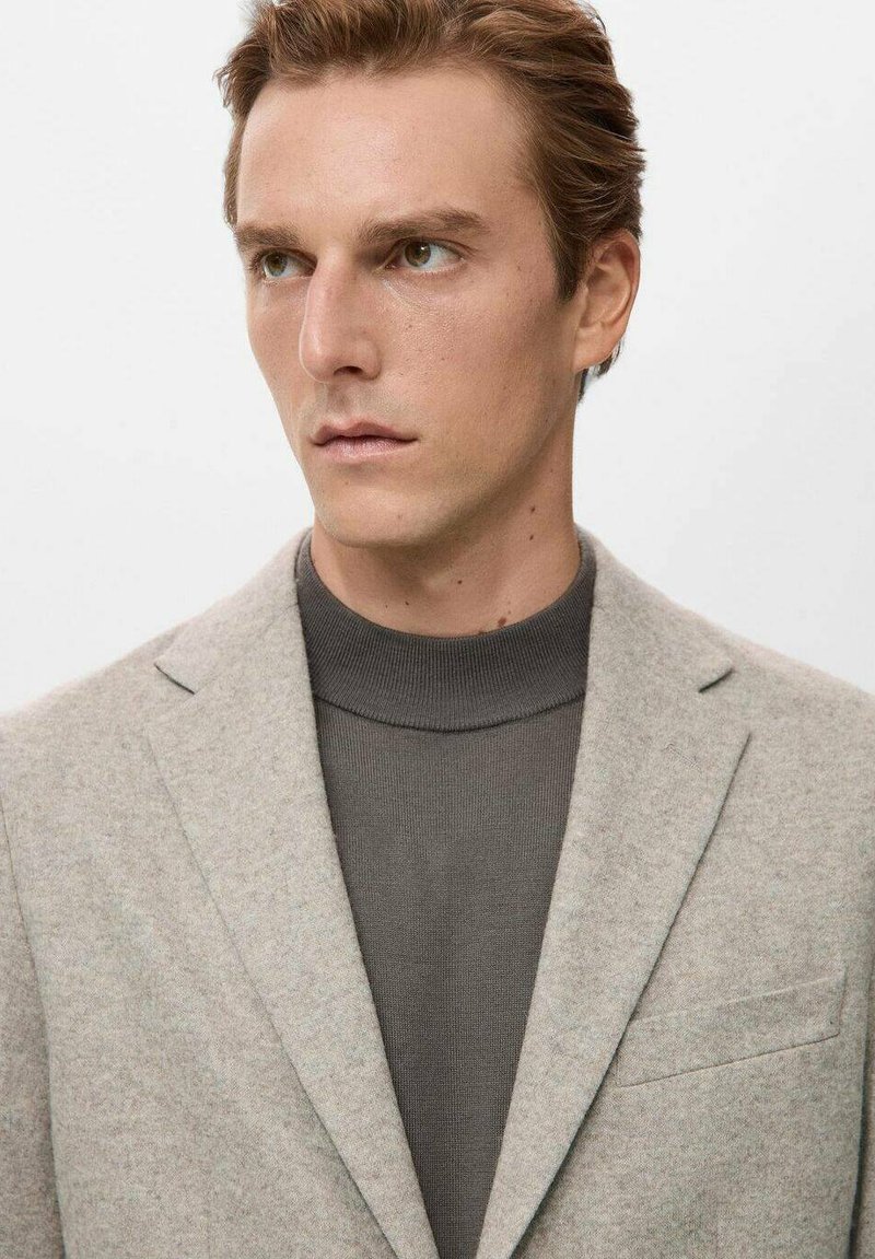 Hellgrauer Blazer mit strukturierter Oberfläche, kombiniert mit einem dunkelgrauen, gerippten Rollkragenpullover. Verfügt über ein Revers mit Kerbe und eine Brusttasche.