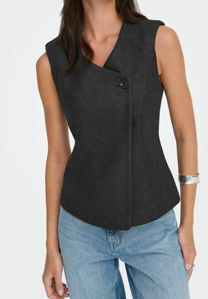 Femme portant un gilet asymétrique sans manches gris foncé boutonné avec un col en V et un jean bleu clair sur fond blanc.