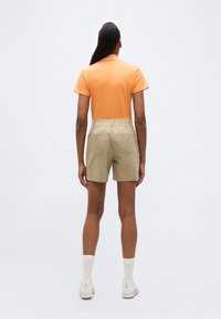 Άτομο που φοράει πορτοκαλί πόλο με κοντά μανίκια, μπεζ σορτς, λευκές κάλτσες και λευκά αθλητικά παπούτσια, στέκεται με την πλάτη προς το μέρος μας σε απλό φόντο.