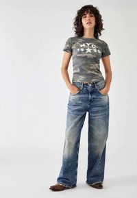 Grijze camouflage t-shirt met witte grafische tekst, gecombineerd met losse lichtblauwe jeans met een vervaagd uiterlijk. Bruine laarzen maken de look compleet.