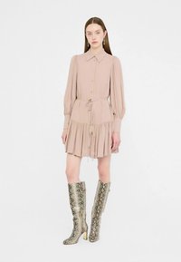 Blassrosa Hemdblusenkleid mit plissiertem Rock, tailliertem Gürtel und langen puffigen Ärmeln. Kombiniert mit kniehohen metallischen Stiefeln mit Schlangenprint.