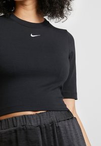 T-shirt noir court fabriqué en tissu extensible, avec un col rond et des manches courtes, arbore un petit logo Nike blanc sur la poitrine.