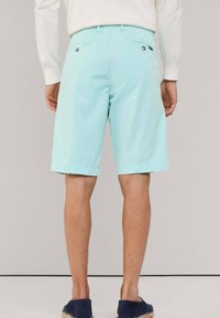 Pantalones cortos de color azul claro con corte recto, que presentan dos bolsillos traseros con botones y un dobladillo doblado. Llevados con zapatos de tela azul marino.