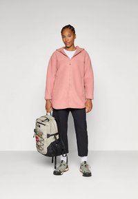 Fleece-Kapuzenjacke in Hellrosa mit Reißverschluss, getragen über einem weißen Hemd. Schwarze Hose und graue Wanderschuhe, trägst einen grauen Rucksack.