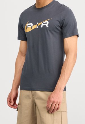Homme portant un t-shirt à manches courtes gris foncé avec le logo Nike swoosh doré et le texte "AIR", assorti à un short cargo beige.