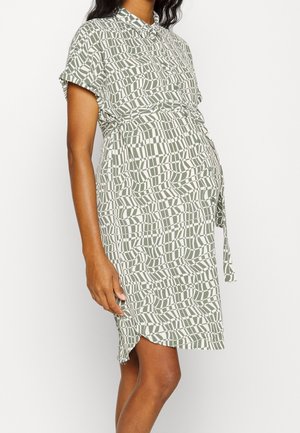 Gravid person iført kortærmet midi-kjole med grønt og hvidt geometrisk cirkelmønster, stående i profil på ensfarvet baggrund.