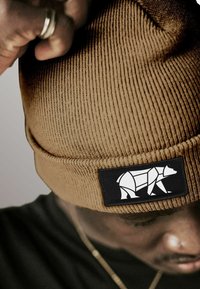 Person wearing a brown knit beanie with a black patch featuring a white geometric bear design, adjusting the hat with a hand.