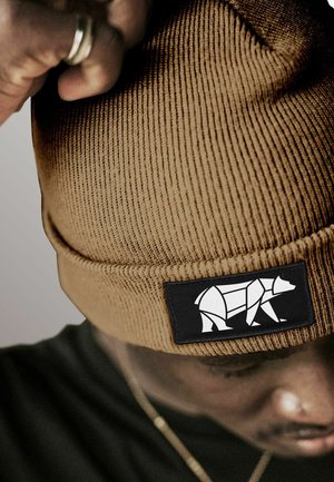 Person wearing a brown knit beanie with a black patch featuring a white geometric bear design, adjusting the hat with a hand.