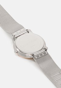 Montre en acier inoxydable argenté avec un bracelet en maille, boîtier rond et dos gravé avec le nom de la marque et les informations sur le modèle.