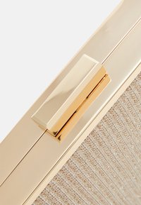 Zlatě tónované zapínání na texturovaném béžovém materiálu. Povrch má jemný třpyt, který zvyšuje celkový elegantní vzhled.