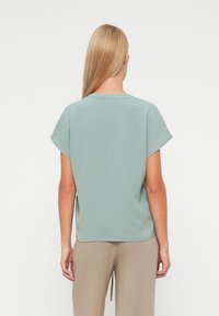 Personne blonde portant un haut texturé vert clair à manches courtes et un pantalon beige, debout, dos tourné sur un fond uni.