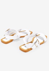 Sandalias planas blancas con tiras cruzadas, diseño de punta cuadrada, suela marrón y un acento de hebilla metálica en la correa del tobillo.