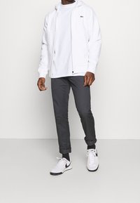 Witte hoodie met rits over een wit T-shirt, gecombineerd met donkergrijze taps toelopende broek en witte sneakers met zwarte accenten.