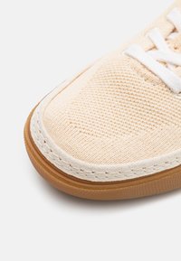 Beige canvas sneakers med en texturerad ovandel, vita snörningar och en gummisul som har sydda detaljer längs kanten.