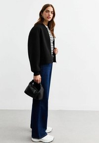 Giacca bomber nera cropped, camicia a righe, jeans blu a gamba larga, sneakers bianche e una piccola borsa nera con una texture morbida e manici arrotondati.