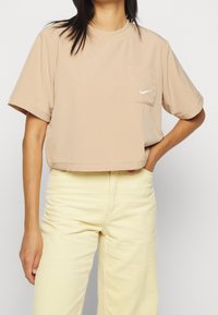 Kvinna iklädd en beige kortärmad t-shirt med en liten Nike-logotyp på fickan och ljusgula manchesterbyxor, hand på höften.