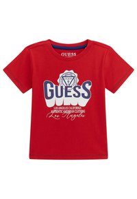 Rød bomulds t-shirt med korte ærmer, der har stor hvid og blå "GUESS" tekst med et cirkulært logo og yderligere hvid tekstbeskrivelse.